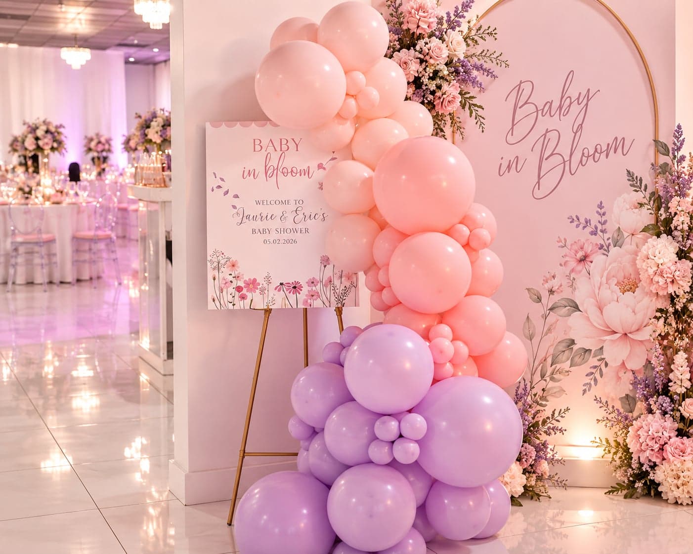 A pink and lavender balloon garland beside a 'Baby in Bloom' welcome sign on a gold easel, in front of a matching floral arch backdrop at a baby shower.